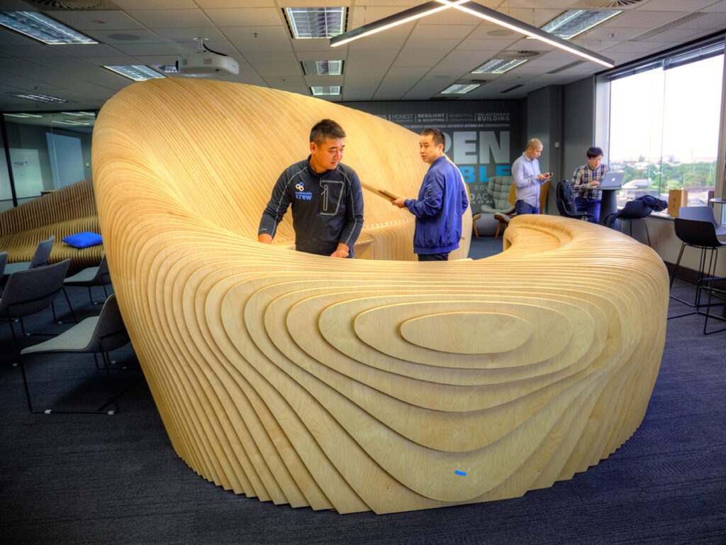 A sleek and modern wooden bench featuring a sculptural form, providing innovative seating in a contemporary office environment. The bench is designed with a flowing design and layered slats, creating a visually appealing and functional centerpiece.