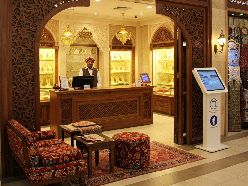 A warm and welcoming gold souk interior with an inviting entrance, including a small reception area with comfortable seating, clear directional signage, and interactive digital elements subtly integrated into the traditional setting. The design reflects the balance between modern convenience and classic market charm.